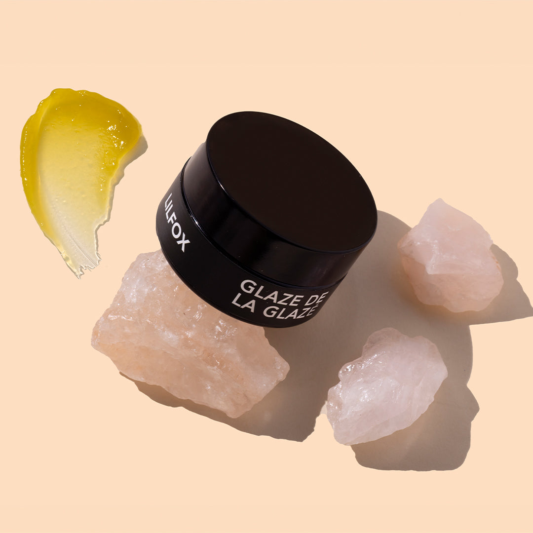 LILFOX | Glaze de la Glaze | Styled Image with Swatch_Sep26 - black jar with a golden yellow balm swatch and pale pink crystals on a peach background