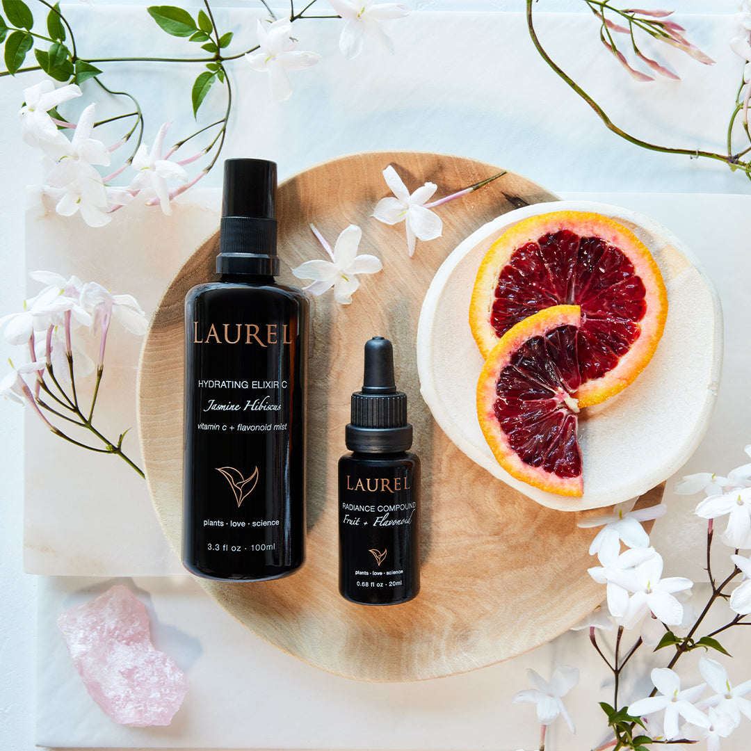 Laurel Skin | Hydrating Elixir C & Radiance Compound | Lifestyle Image - mist and  serum displayed on a wooden tray with blood orange slices, white flowers, and a rose quartz crystal