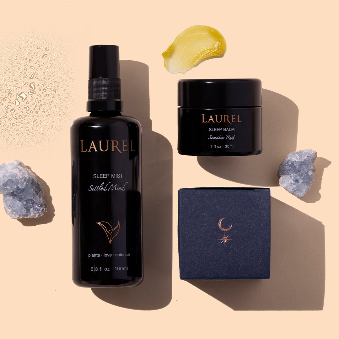 Laurel Skin | Sleep Set | Styled Image with Swatch_Sep26 - Laurel Sleep Collection featuring black Sleep Mist bottle, Sleep Balm jar, navy boxes, yellow balm swatch, water droplets, and crystals on a peach background