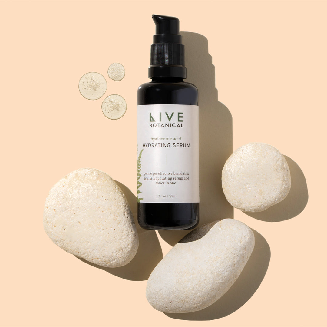 Live Botanical | Hyaluronic Acid Serum | Styled Image with Swatch_Sep26 - serum bottle with cream swatches and white stones on a peach background