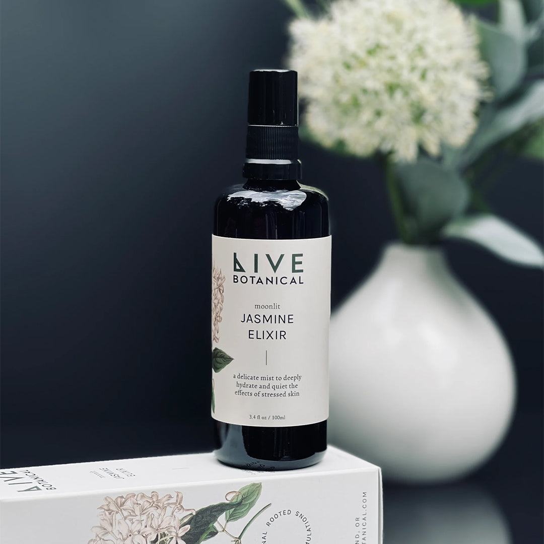 Live Botanical | Moonlit Jasmine Elixir | Lifestyle Image - dark bottle on its product packaging beside a vase of white flowers in a softly lit setting