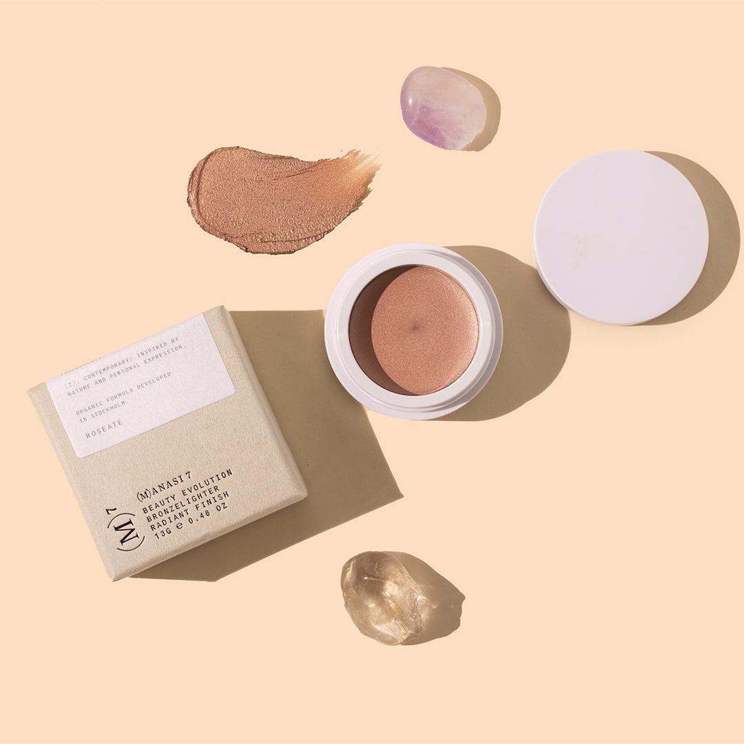 Manasi 7 | Bronzelighter Roseate | Styled Image with Swatch_Oct26 - warm bronze-peach highlighter with a matching metallic swatch, white packaging, beige box, and pale purple and clear crystals on a warm peach background
