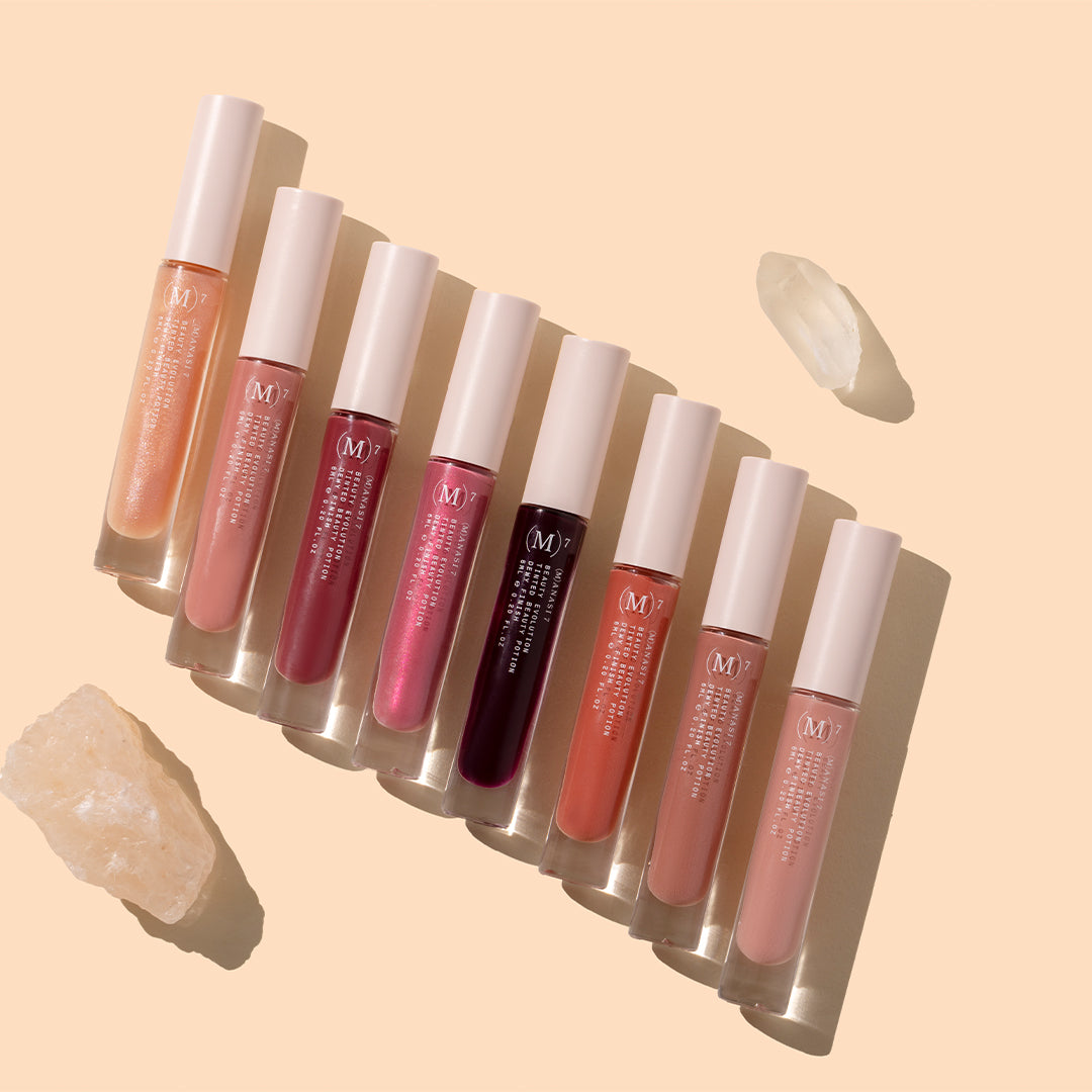 Manasi 7 | Tinted Beauty Potion | Styled Image with Swatch_Oct26 - Eight glosses in sheer peach, dusty rose, berry red, vibrant pink, deep burgundy, coral, rosy nude, and pale pink, with white caps and crystals on a warm peach background