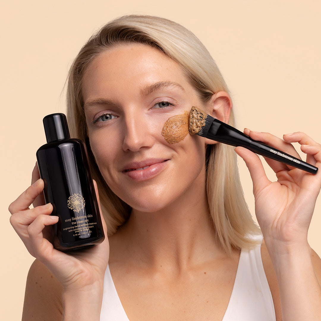 May Lindstrom Skin | The Clean Dirt | Model Image with swatch - person holding black bottle near face also holding a brush applying mask to skin, on peach background