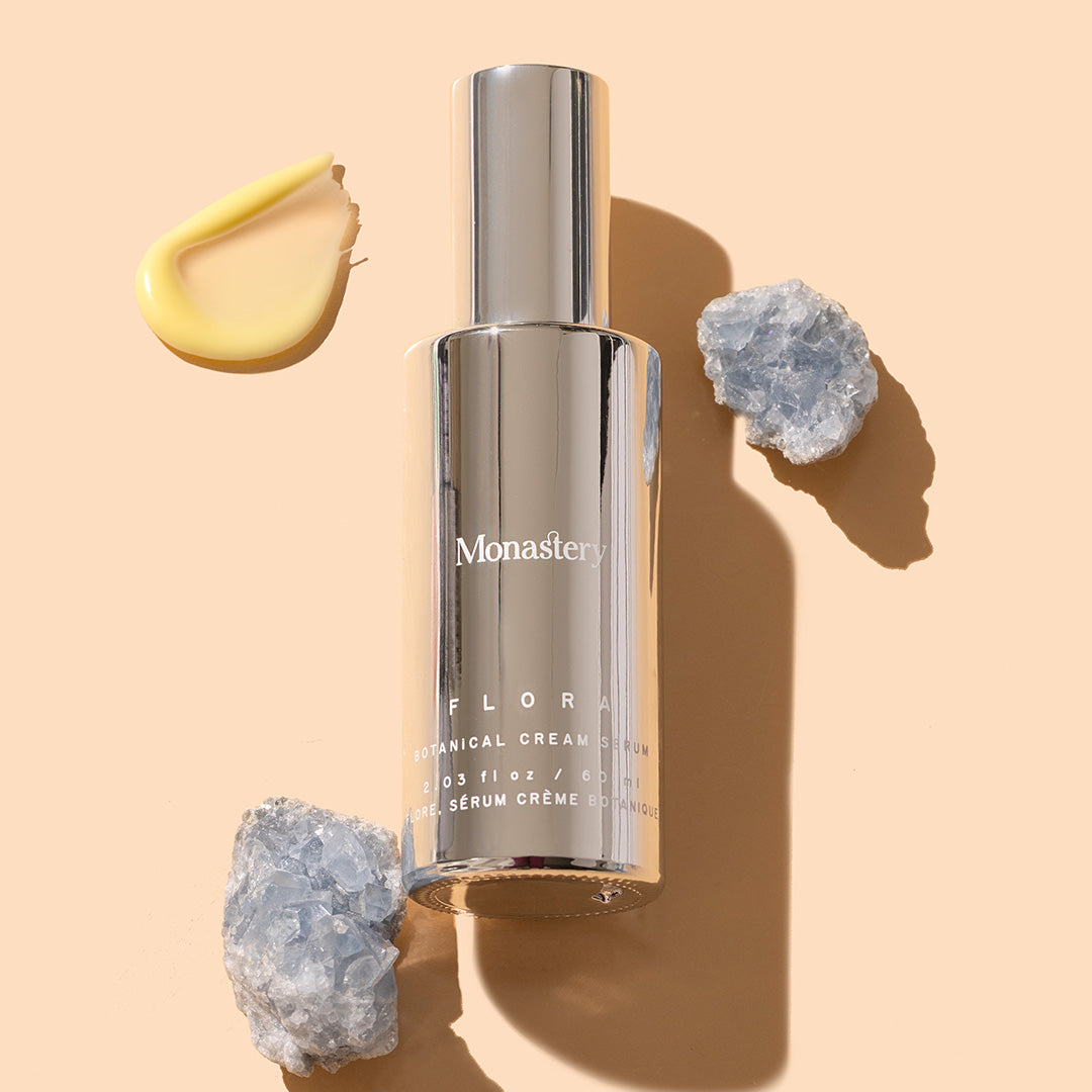 Monastery | Flora Botanical Cream Serum | Styled image with swatch, silver bottle with silver cap on peach background with yellow product cream swatch and two blue crystals