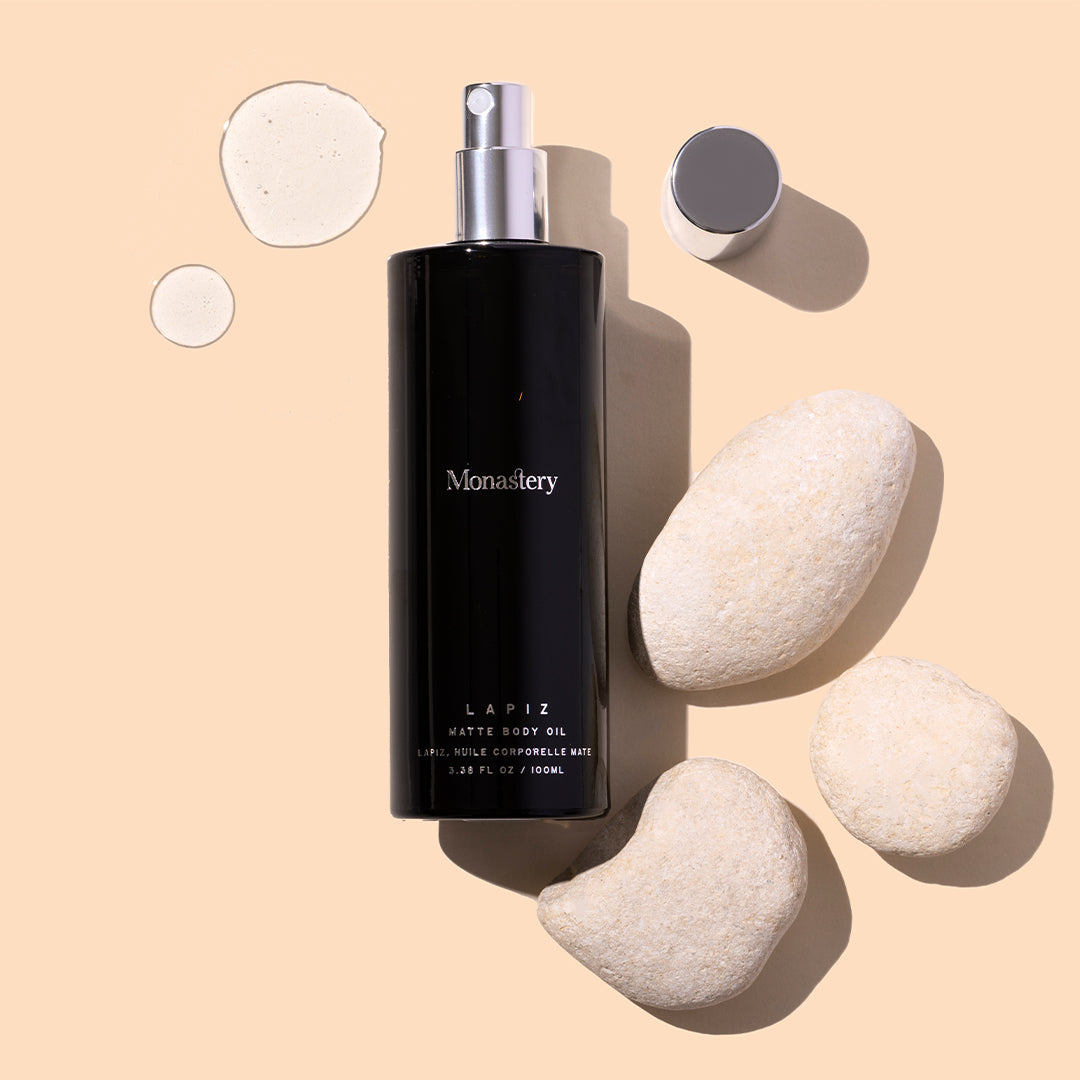 Monastery | Lapiz Matte Body Oil | Styled Image with Swatch_Jul26 - black bottle with oil swatches and white stones on a beige background