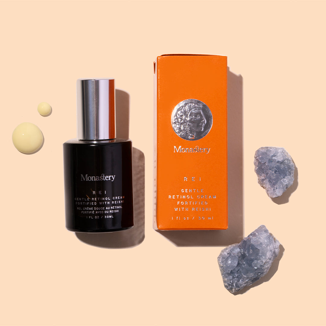 Monastery | REI Gentle Retinol Cream | Styled Image with Swatch_Jul26 - black bottle with orange box, cream droplets, and blue crystals on a beige background