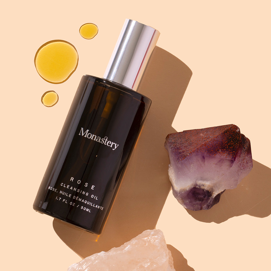 Monastery | Rose Cleansing Oil | Styled Image with swatch, dark bottle with silver cap on peach background with yellow oil swatch and crystals