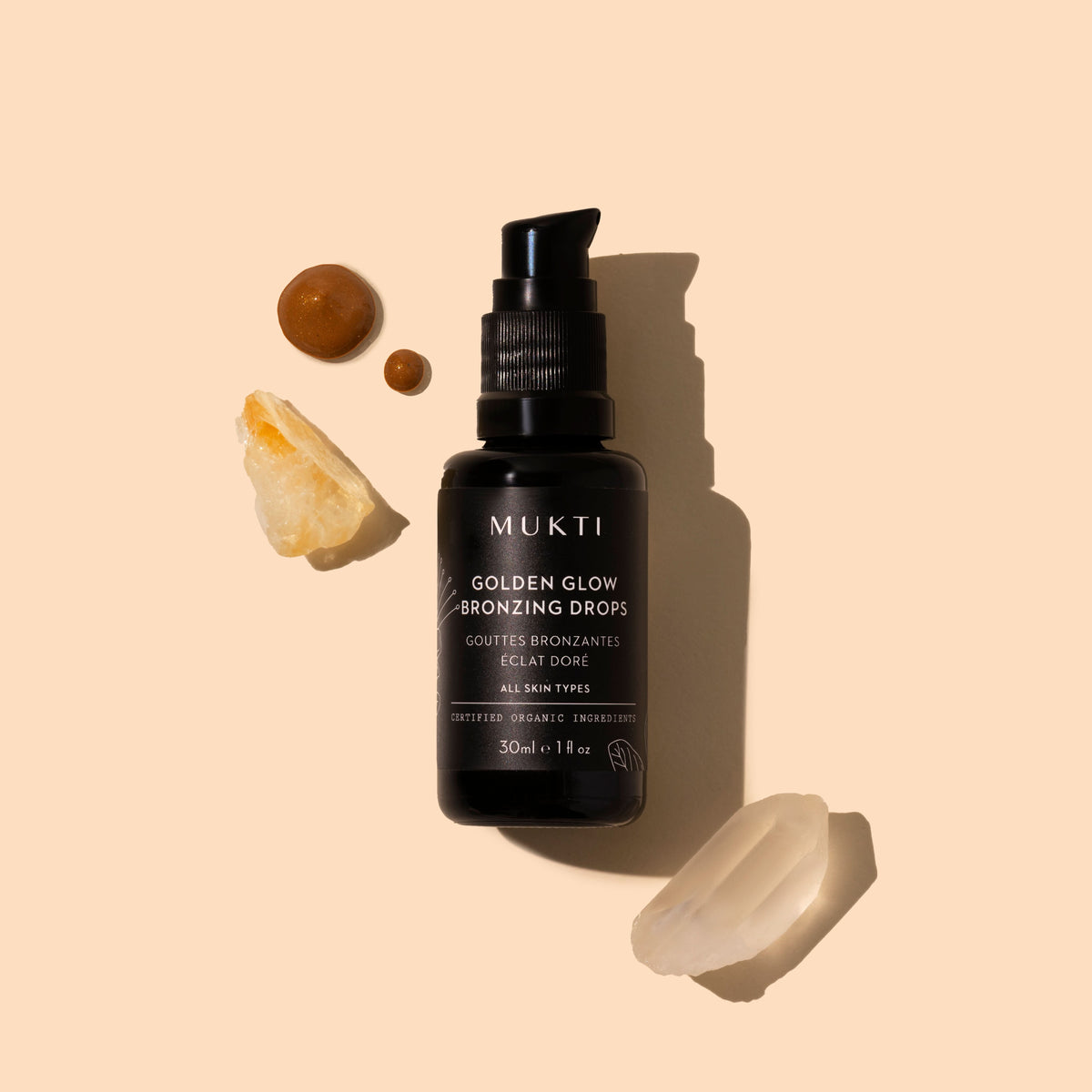 Mukti | Golden Glow Bronzing Drops | Styled Image with swatch | A bottle of MUKTI Golden Glow Bronzing Drops centered on a warm beige background, styled with natural ingredients including a slice of orange, a small brown seed, and a translucent crystal. The bottle is black with a black pump dispenser, and the text on the bottle is white.