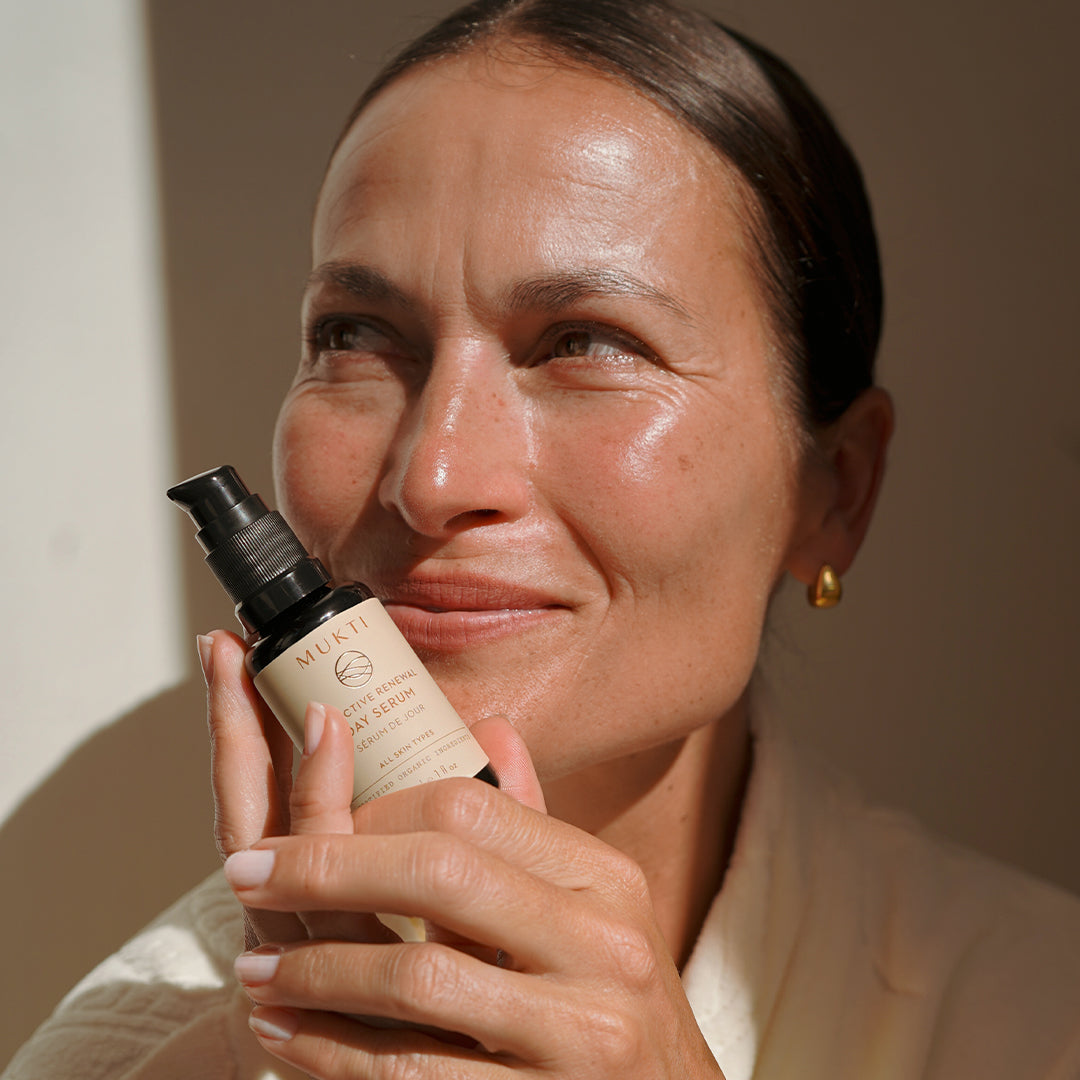 Mukti Organics | Bioactive Renewal Day Serum | Model Image - woman holding black pump bottle in warm sunlight against a neutral brown background