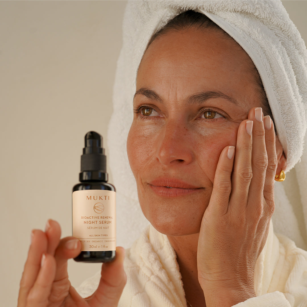 Mukti Organics | Bioactive Renewal Night Serum | Model Image - woman in a white towel holding black pump bottle against a warm beige background