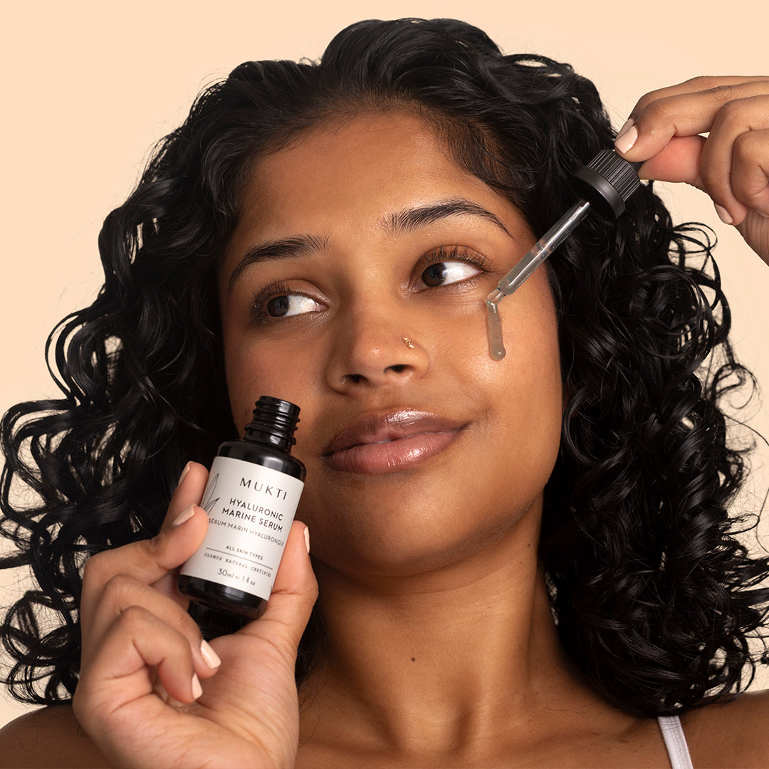 Mukti Organics | Hyaluronic Marine Serum | Model Image - person applying serum with a glass dropper while holding the serum bottle against a neutral background