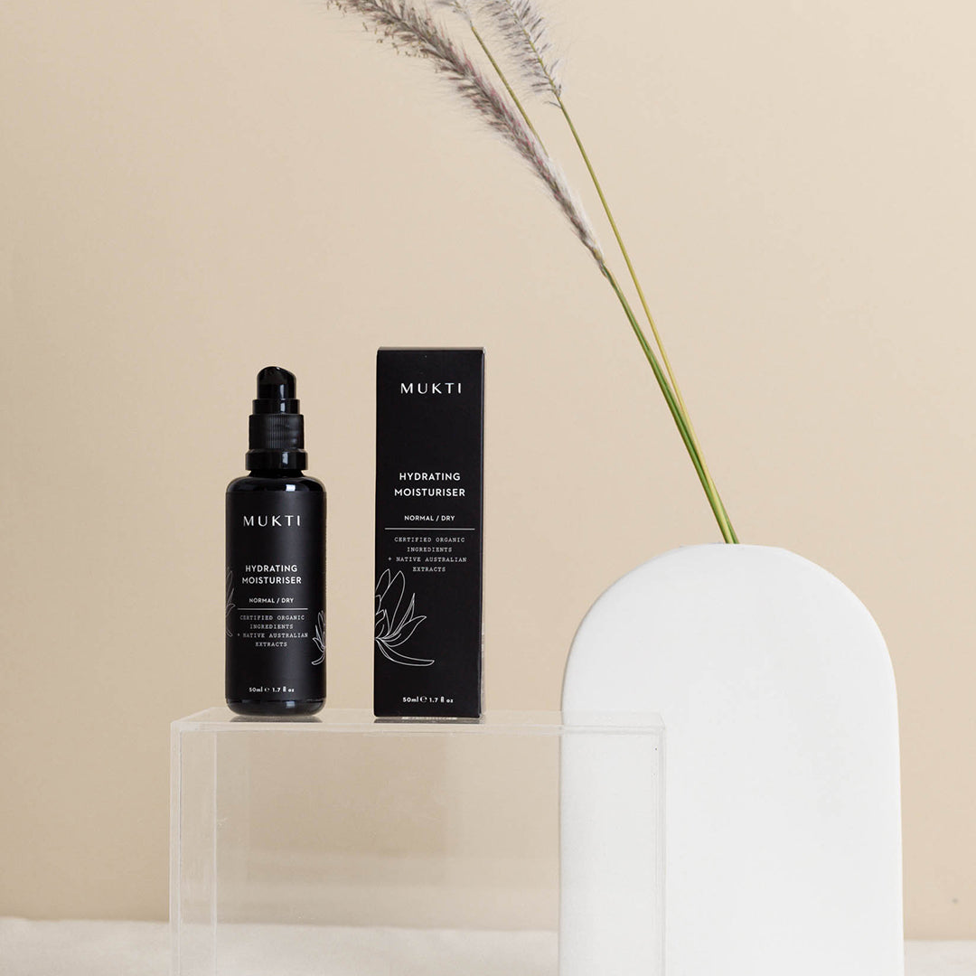 Mukti Organics | Hydrating Moisturiser | Lifestyle Image - black bottle and box displayed on a clear stand against a beige background