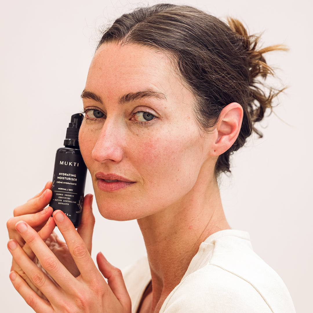 Mukti Organics | Hydrating Moisturiser | Model Image - woman holding black moisturiser bottle beside her face against a white background