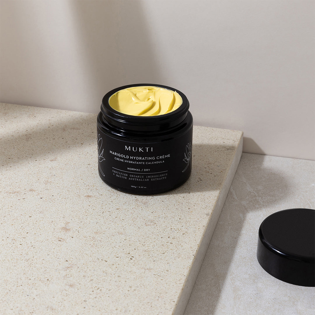 Mukti Organics | Marigold Hydrating Crème | Lifestyle Image - open black jar showing yellow cream on a beige stone surface
