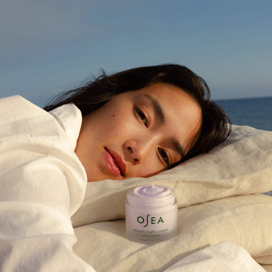 OSEA | Dream Night Cream with Bio-Retinol | Model Image - person resting on white pillows beside an open jar of cream