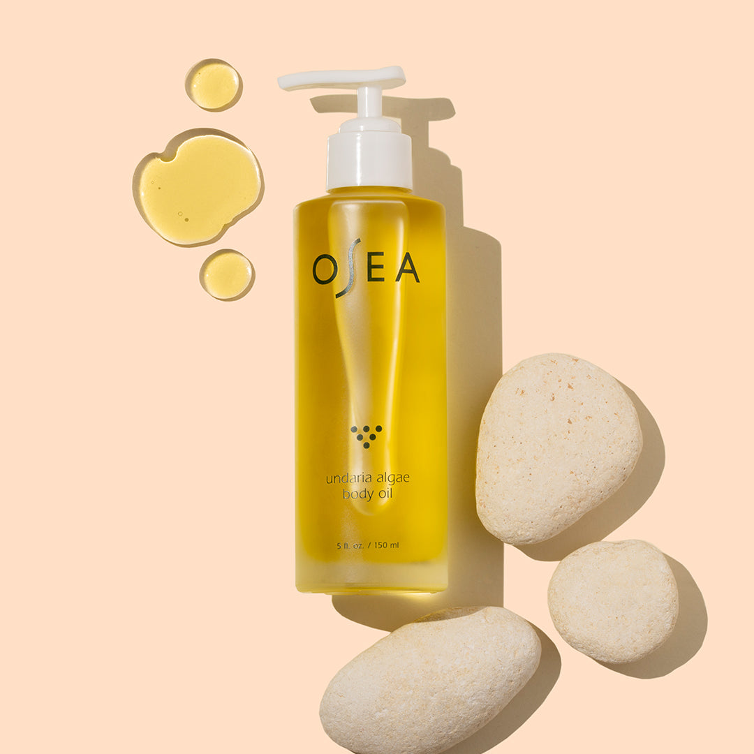 OSEA | Undaria Algae Body Oil | Styled Image with Swatch_Sep26 - body oil bottle with golden yellow oil swatch on a peach background with white stones
