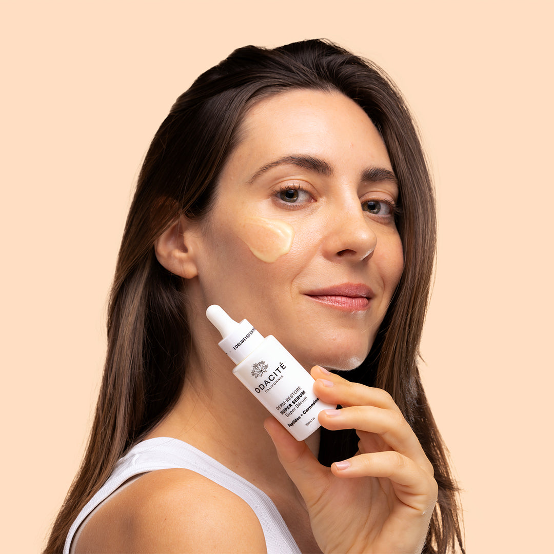 Odacité | Edelweiss Extrême Derm-Restore Super Serum | Model Image with Swatch - woman holding white bottle with swatch on her cheek against a beige background