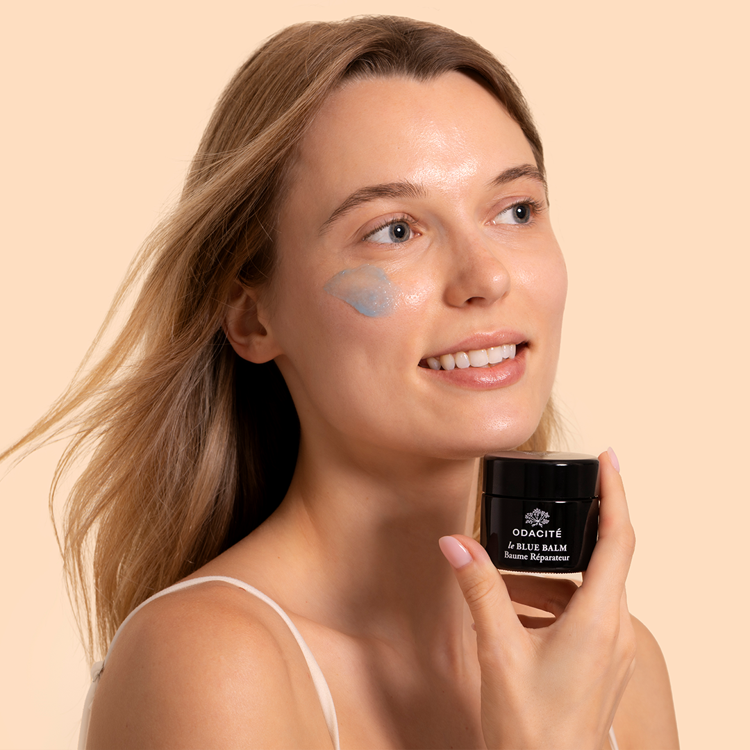 Odacité | le Blue Balm | Model Image - woman holding a jar of balm while wearing a swipe of the product on her cheek against a beige background
