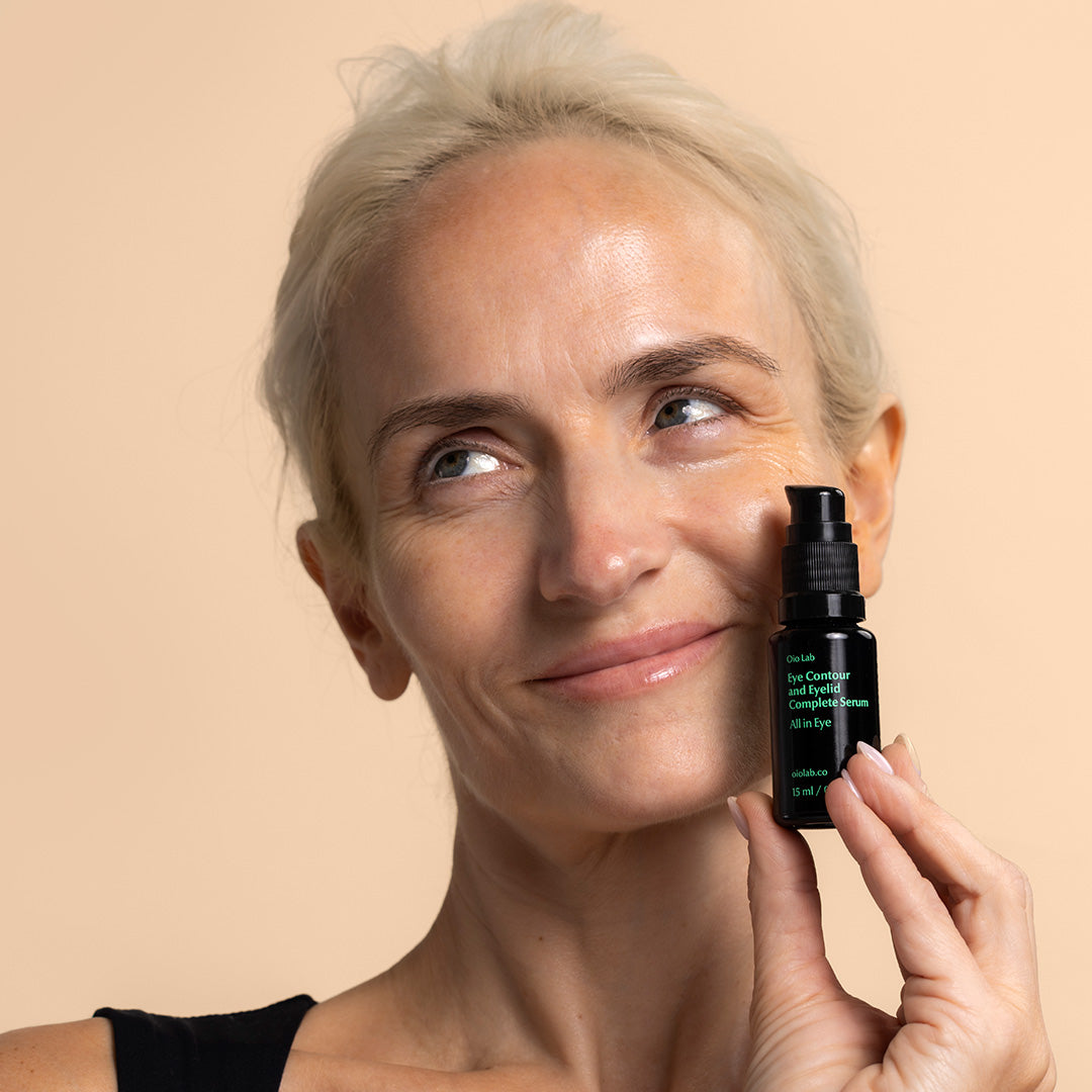 Oio Lab | All In Eye Contour and Eyelid Complete Serum | Model Image, model holding black jar with green writing near face with product applied to under eye