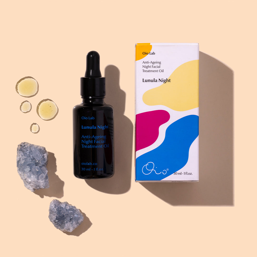 Oio Lab | Lunula Night Facial Oil | Styled Image with Swatch_Aug26 - black bottle with product box, oil droplets, and crystal accents on a beige background