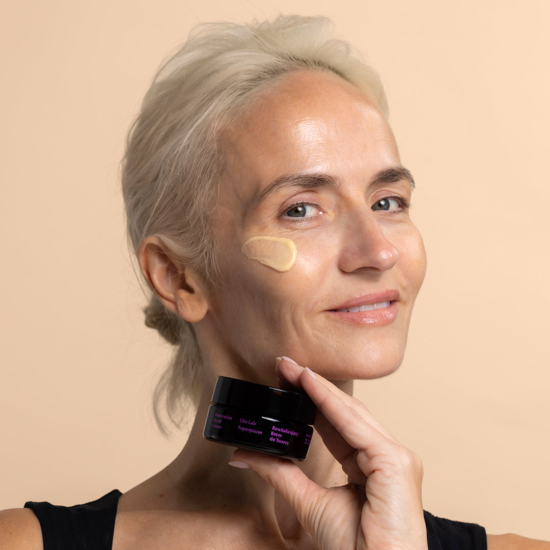Oio Lab | Superpause Restorative Facial Cream | Model Image - woman holding black cream jar with cream applied to cheek against a beige background.
