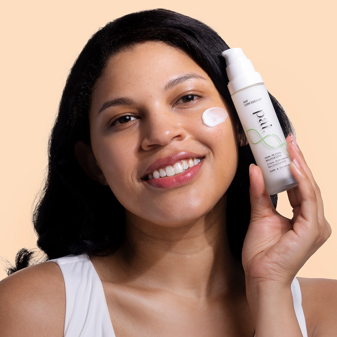 Pai | PDRN Revival Moisturizer | model image | A woman with dark hair holds a white bottle of Dr. G's Radiance Skin Tint against a solid beige background, with a dollop of the tinted cream shown on her cheek.