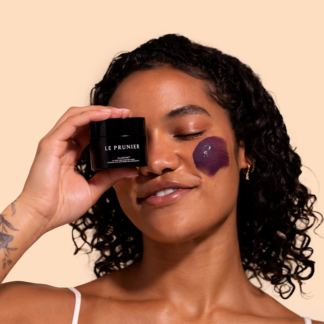 Plumtox | Clarifying Enzyme Mask | Model Image_Jul26 - model holding mask jar with a purple mask applied to the cheek against a beige background