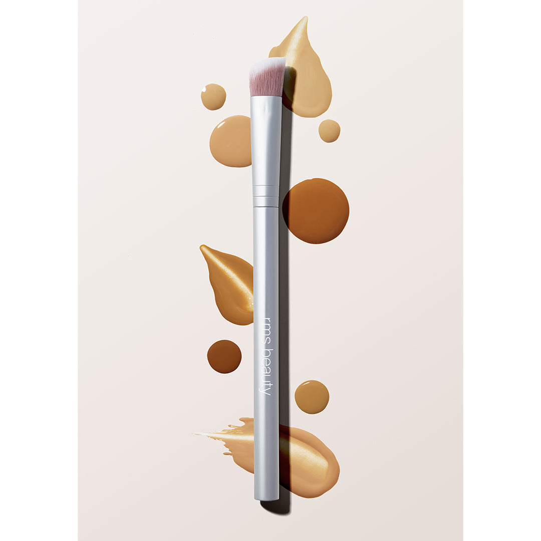 RMS Beauty | Concealer Brush | Lifestyle Image - brush with soft pink-and-white bristles and a matte silver handle displayed with cream complexion product swatches in varying shades