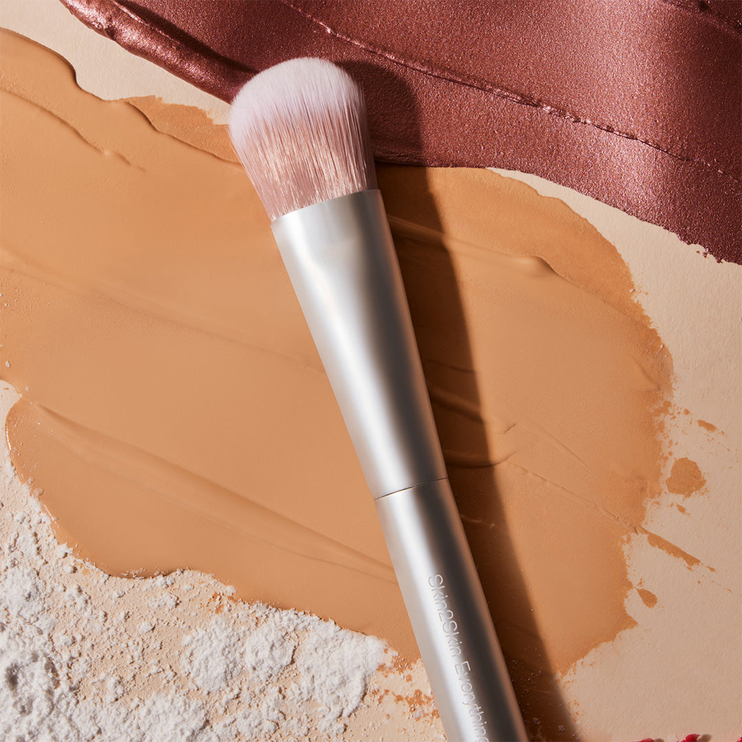 RMS Beauty | Everything Brush | Flatlay Image - brush resting on cream, powder, and bronze makeup textures