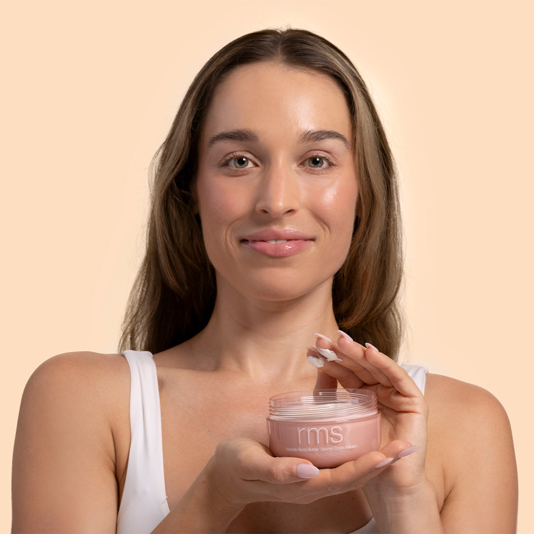 RMS | Beauty Kakadu Body Butter | Model Image_Aug26 - model holding an open pink jar with a small amount of cream on their fingertips against a soft peach background