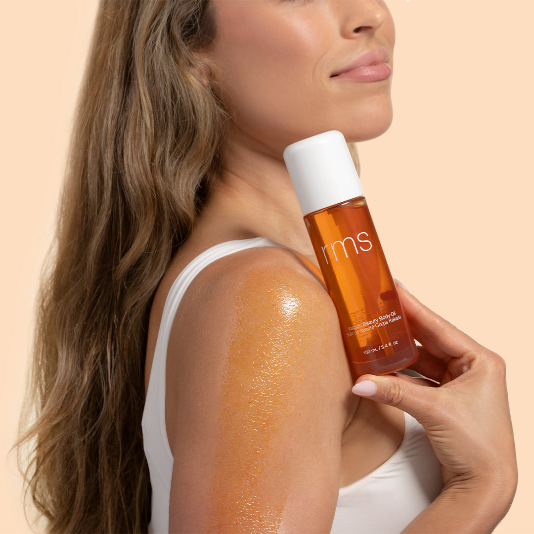 RMS Beauty | Kakadu Body Oil | Model Image_Aug26 - model holding bottle beside a glowing, oil-coated shoulder while wearing a white tank top against a light beige background