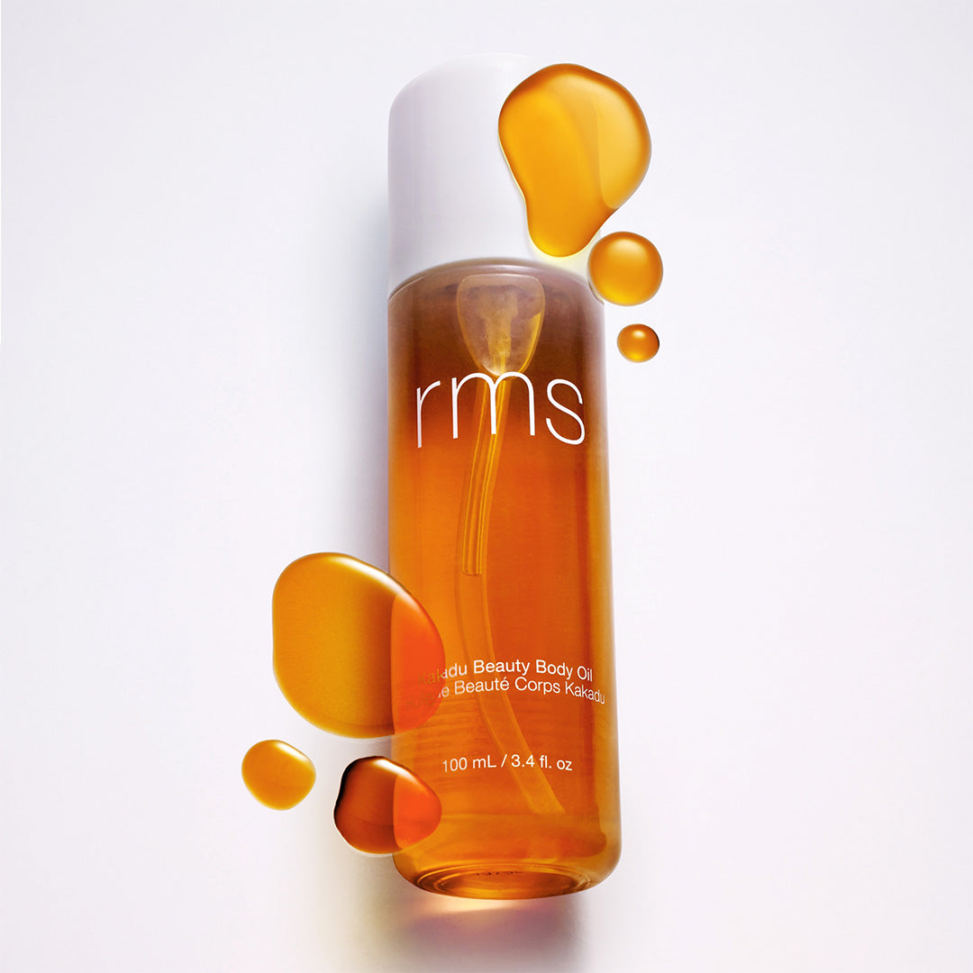 RMS Beauty | Kakadu Body Oil | Product with Swatch Image - bottle surrounded by golden oil drops on a softly lit background