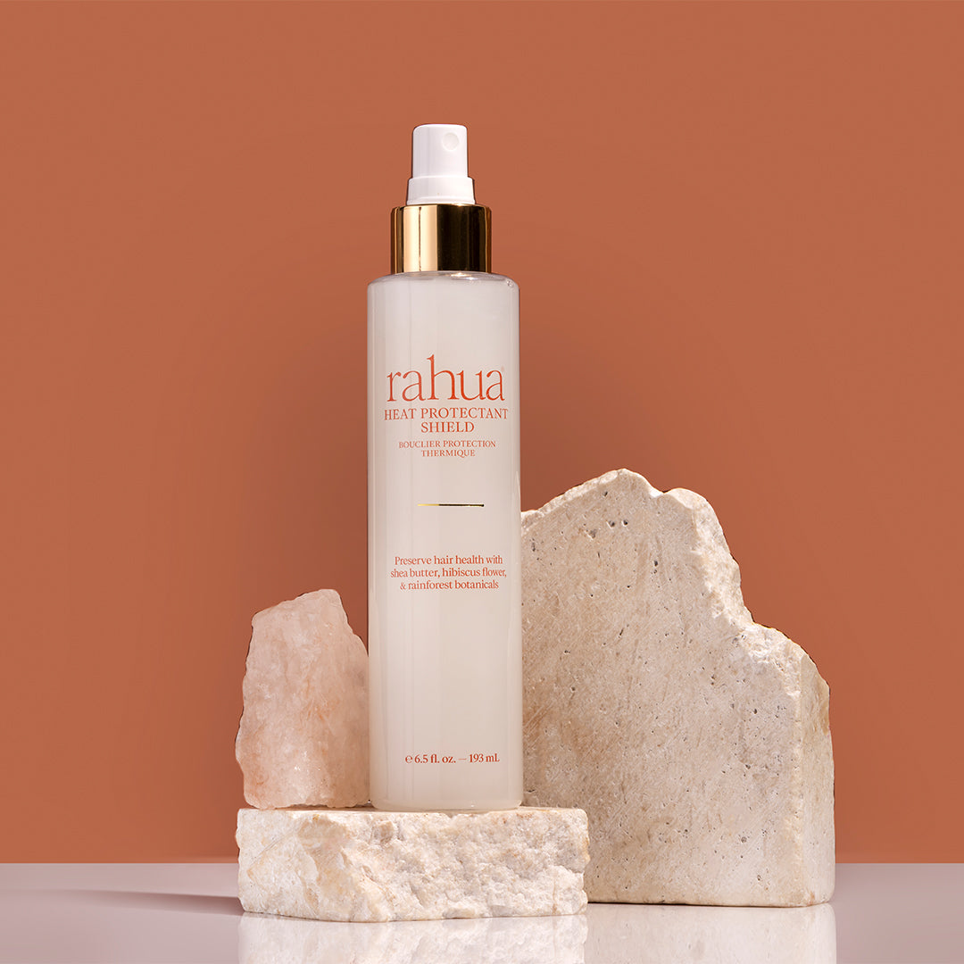 Rahua | Heat Protectant Shield | Product Image2 - spray bottle with white and gold cap with stones and crystals against a brown background