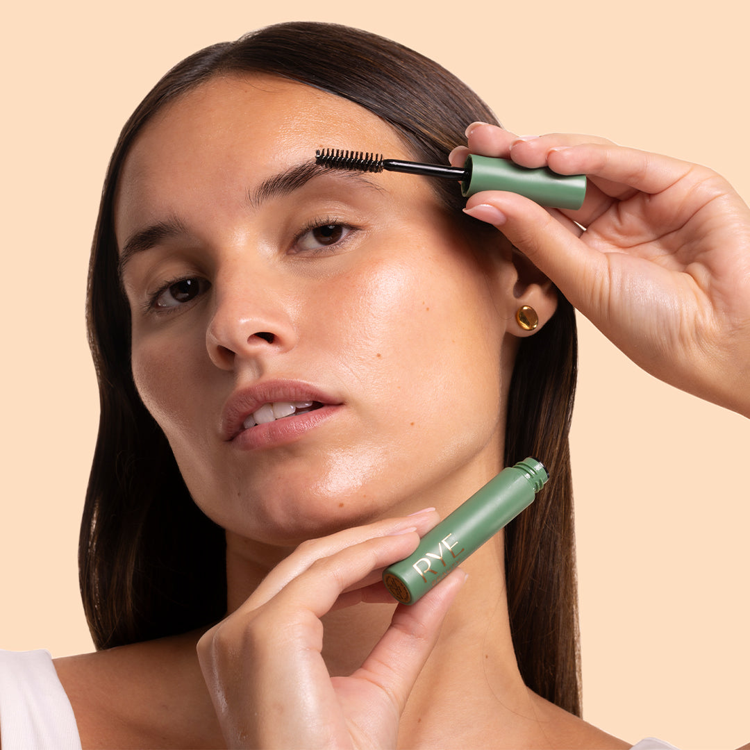 Rye Beauty | Brow Grooming Serum | Model Image - model applying brow serum to her eyebrow while holding the green tube against a peach background
