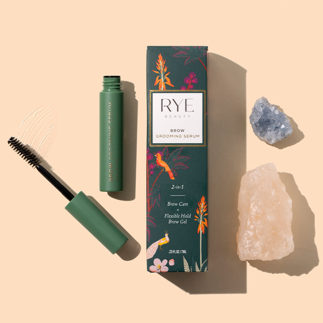 Rye Beauty | Brow Grooming Serum | Styled Image - green packaging, spoolie applicator, clear swatch, and crystals on a peach background