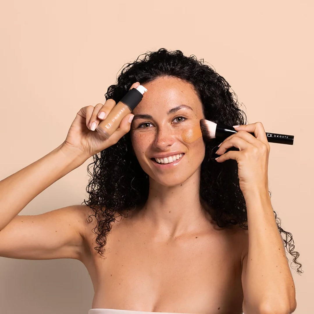 TOK Beauty | Foundation Brush | Model Image - smiling model applying foundation with brush while holding a foundation bottle against a beige background