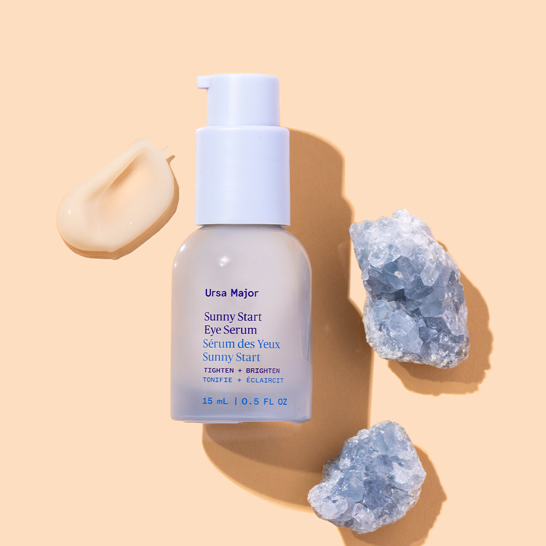 Ursa Major | Sunny Start Eye Serum | Styled Image with Swatch - bottle displayed with a cream swatch and two blue crystals on a beige background