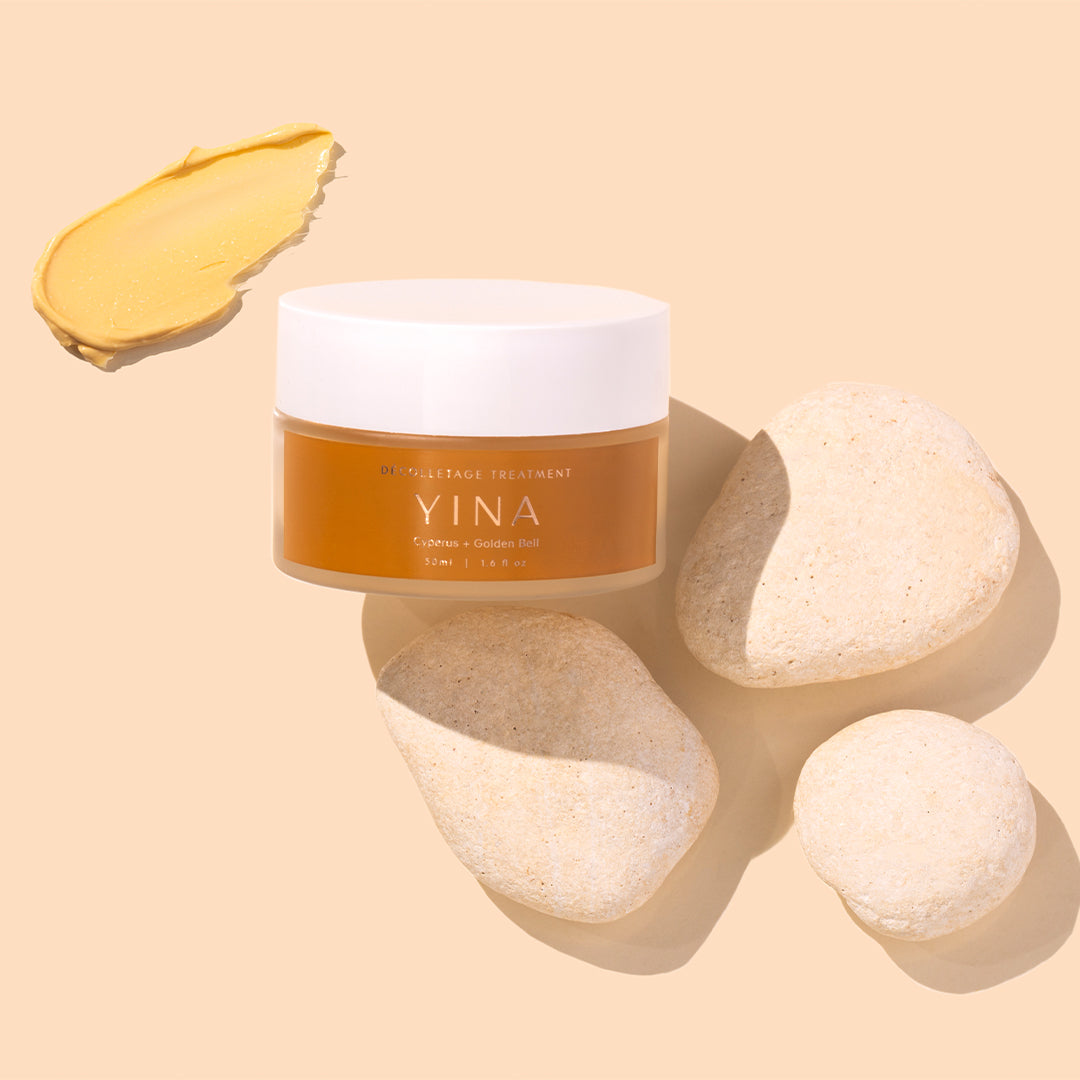 YINA | Décolletage Treatment | Styled Image with Swatch_Aug26 - amber jar with a white lid, displayed beside smooth white stones and a golden cream swatch on a soft beige background