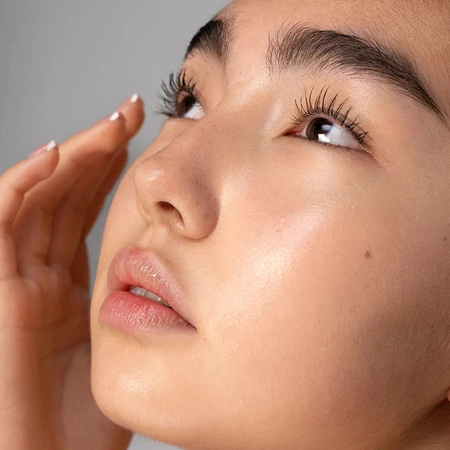 RMS Beauty | Straight Up Mascara | Close-up of a woman's face with long, dark, and curled eyelashes, looking upward, with her hand near her face against a neutral gray background.