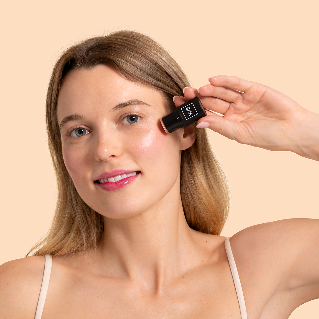 withSimplicity | Tri-Stick | Model Image - woman applying a blush stick directly to her cheek against a beige background